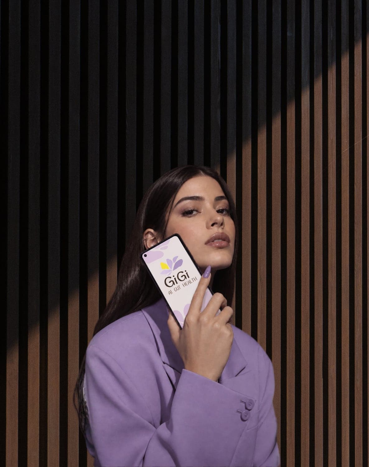 A woman in a lilac suit holding a phone with the Gigi app, against a dark striped wall